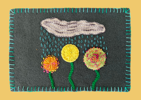 Photo of a mug rug on a yellow background with wool appliqué cloud and flower shapes embellished with perle cotton.