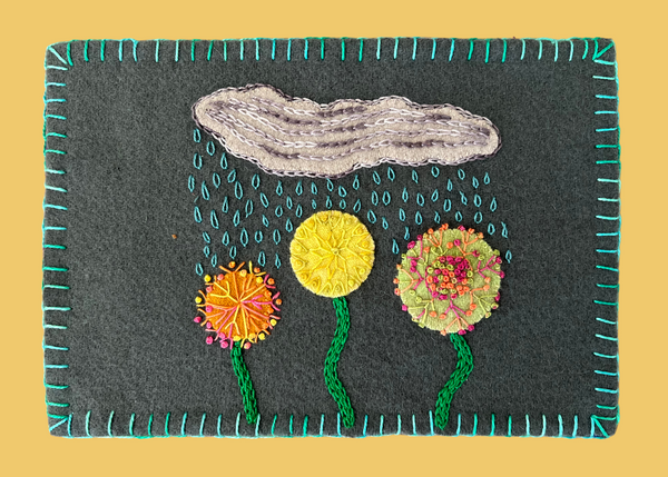 Photo of a wool mug rug on a yellow background with wool appliqué clouds and flowers and perle cotton embroidery stitches.