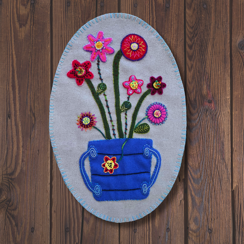 Photo of a wool appliqué oval mat with wool bucket and flowers embellished with perle cotton embroidery stitches on a dark wook background.