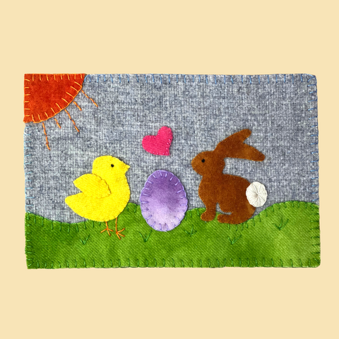 A photo of a wool applique bunny and chick on a grey wool background with green grass and hand embroidery stitches